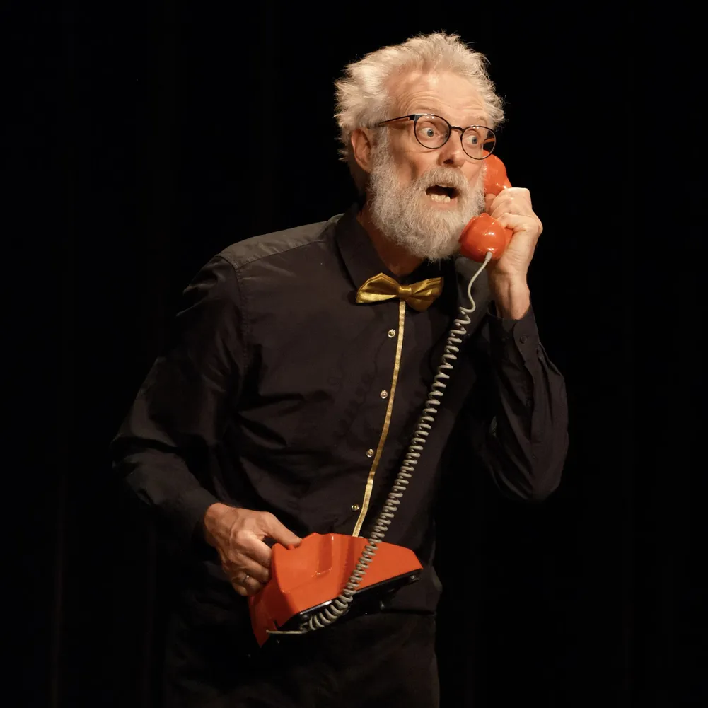 Close-up van een koorlid (tenor) met een baard, die tijdens een performance expressief telefoneert met een klassieke rode hoorn.